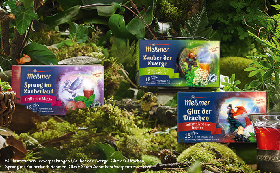 Zwei Schachteln Meßmer-Tee mit fantasievollen Motiven stehen in einer üppigen, grünen Waldlandschaft neben einer Glastasse mit leuchtend rotem Tee. Moos und Blätter umgeben die Szene.