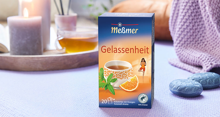 Eine Schachtel Meßmer Gelassenheit-Tee steht auf einer hellvioletten Fläche mit Kerzen, einer Tasse Tee, Dekosteinen und einer Strickdecke im Hintergrund.