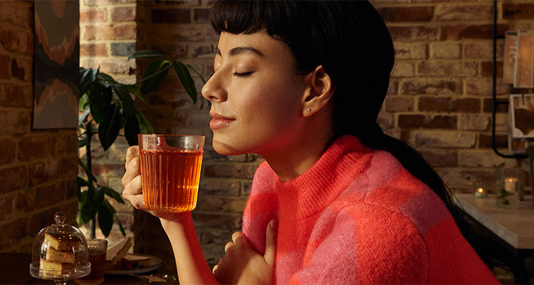 Eine Frau in einem rosafarbenen Pullover sitzt mit geschlossenen Augen und einem Lächeln an einem Tisch, hält eine Glastasse mit Tee an ihr Gesicht und genießt das Aroma in einem gemütlichen Raum mit freiliegenden Ziegelwänden.