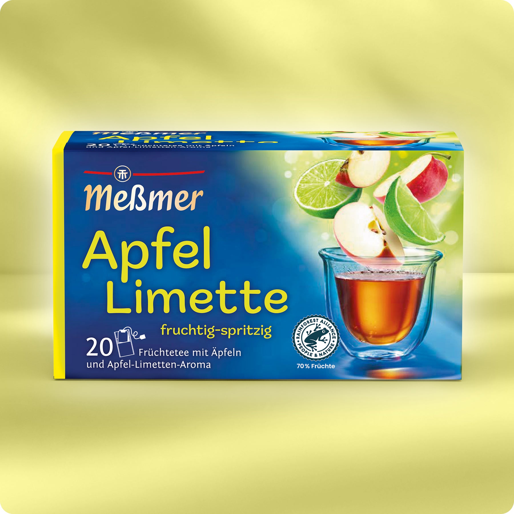 Eine Schachtel Meßmer DE Apfel-Limette Tee zeigt Äpfel und Limettenscheiben über einem Glas Tee. Die blau-gelbe Verpackung zeigt Fruchtabbildungen und Text auf hellgelbem Hintergrund.