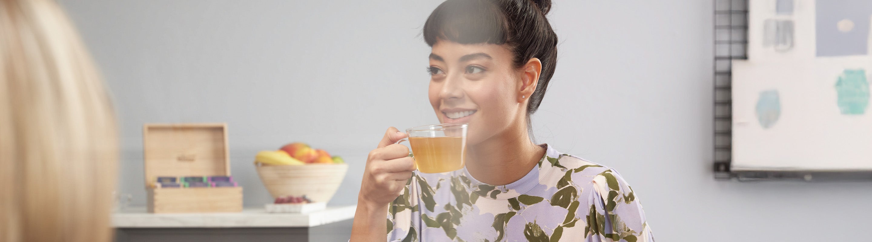 Eine Frau mit dunklem Haar in einem Dutt lächelt, während sie eine Glastasse mit Tee hält. Sie trägt ein geblümtes Hemd und sitzt in einem Haus, mit einer Obstschale und einer hölzernen Teedose im Hintergrund.