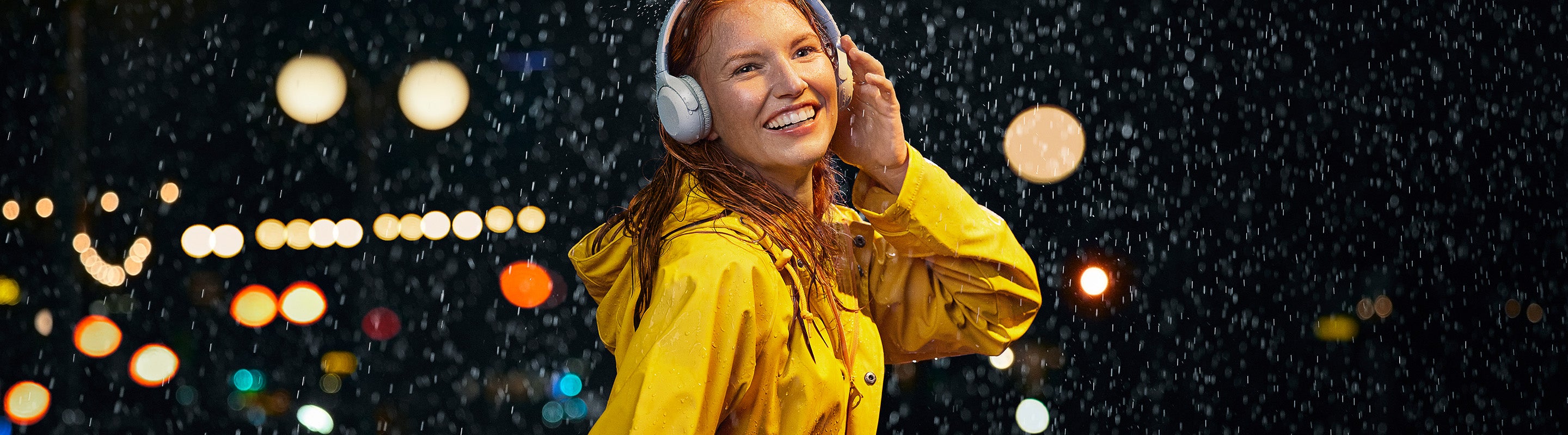 Eine lächelnde Frau in einem gelben Regenmantel hört Kopfhörer und genießt den nächtlichen Regen, mit verschwommenen Lichtern der Stadt im Hintergrund.