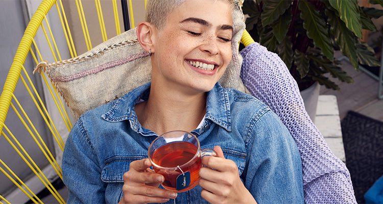 Eine Person mit kurzen blonden Haaren, die ein Jeanshemd trägt, sitzt in einem gelben Stuhl, lächelt mit geschlossenen Augen und hält eine Glastasse mit Tee. Hinter ihr liegen ein Kissen und eine lila Strickdecke.