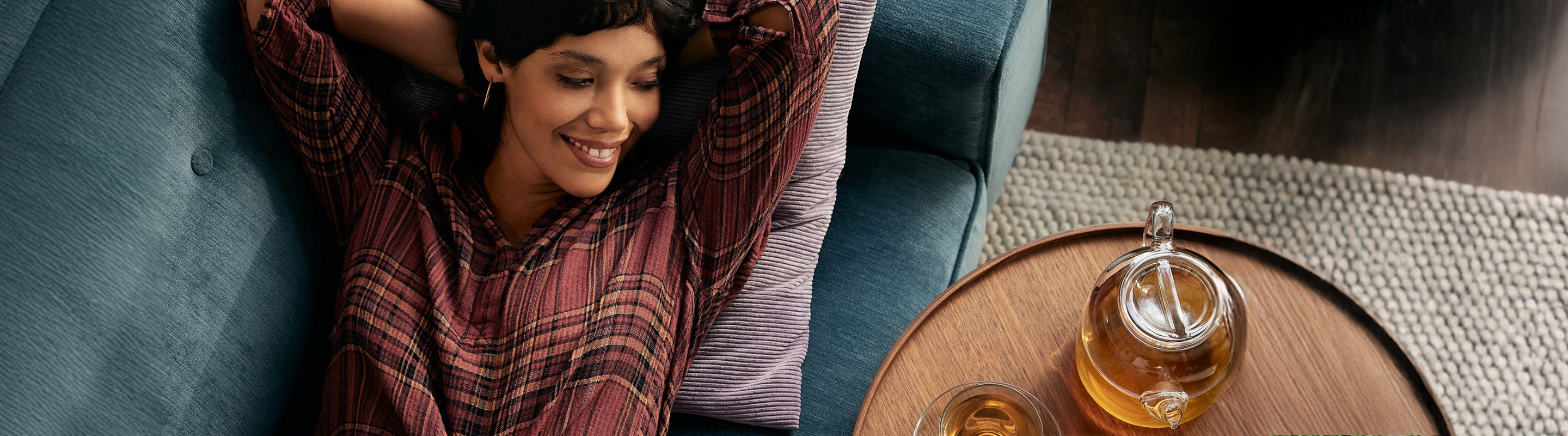 Eine Frau in einem gestreiften Hemd entspannt sich auf einer blauen Couch und verschränkt lächelnd die Arme hinter dem Kopf. Eine gläserne Teekanne und eine Tasse Tee stehen auf einem runden Holztisch in der Nähe.