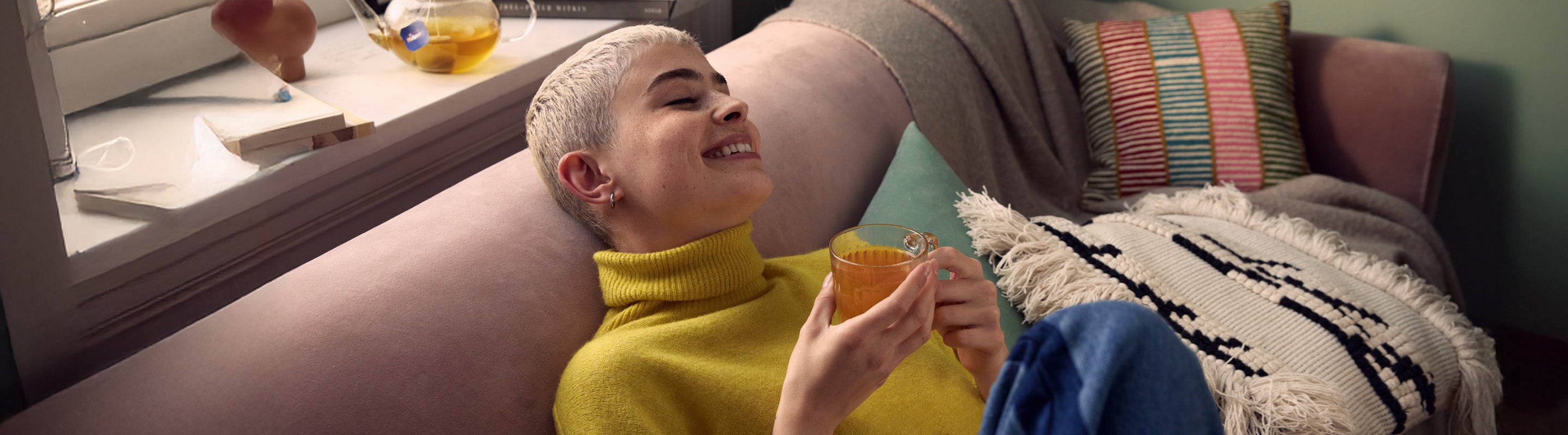 Eine Person mit kurzen blonden Haaren, die einen gelben Pullover trägt, entspannt sich auf einem Sofa, während sie lächelt und eine Tasse Tee in der Hand hält. Neben ihr liegen Kissen und eine Decke und im Hintergrund steht eine Teekanne auf der Fensterbank.