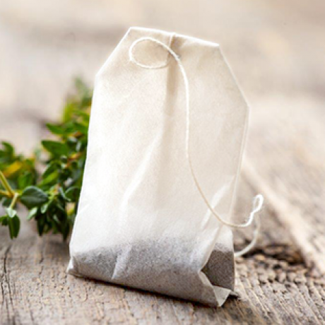 Nahaufnahme eines Teebeutels mit einer Schnur, der auf einer hölzernen Oberfläche liegt. Im Hintergrund ist ein grünes, blättriges Kraut unscharf zu erkennen.