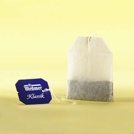 Ein Meßmer Klassik Teebeutel mit blauem Etikett und weißer Schnur ist aufrecht vor gelbem Hintergrund abgebildet. Der Teebeutel enthält in der unteren Hälfte dunkle Teeblätter.