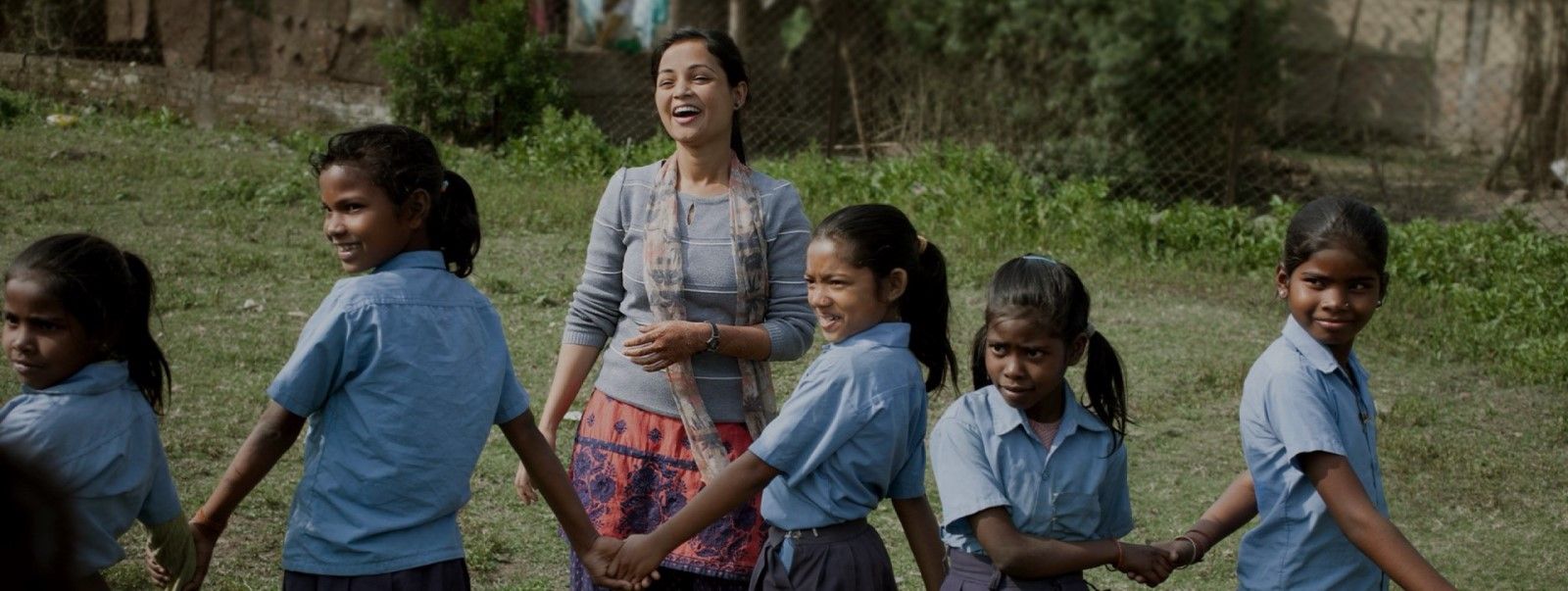 Eine lächelnde Frau steht draußen mit einer Gruppe junger Mädchen in blauen Uniformen, die sich im Kreis an den Händen halten und gemeinsam auf einer Wiese mit Bäumen und einem Zaun im Hintergrund spielen.