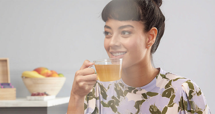 Eine lächelnde Frau mit dunklem Haar in einem Dutt hält eine Glastasse mit Tee. Sie trägt ein blumengemustertes Hemd und sitzt in einem hellen Raum mit einer Obstschale im Hintergrund.