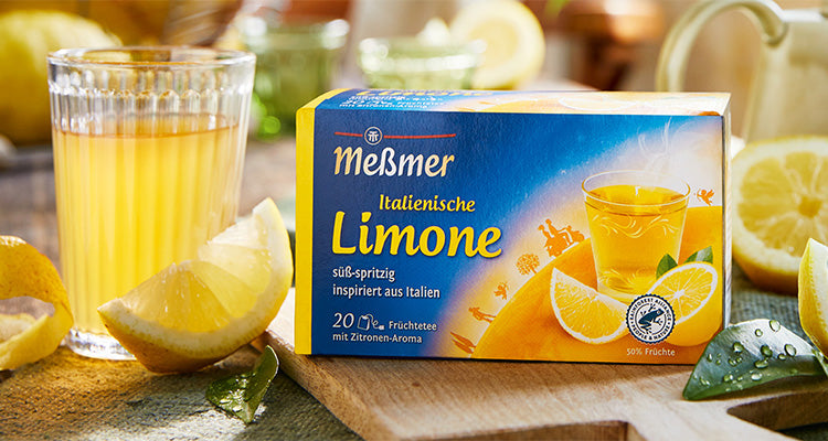 Eine Schachtel Meßmer Italian Lemon Früchtetee steht auf einem Holztisch, daneben ein Glas Zitronentee, frische Zitronenscheiben und ein grünes Tuch, alles in hellem Tageslicht.