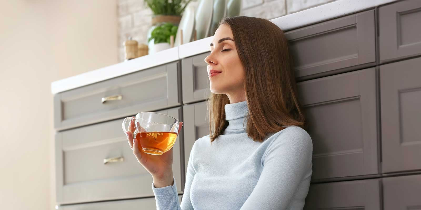 Eine Frau mit lockigem Haar lächelt mit geschlossenen Augen, während sie eine Glastasse mit rotem Tee in der Hand hält. Im Hintergrund ist ein geschmückter Weihnachtsbaum zu sehen.