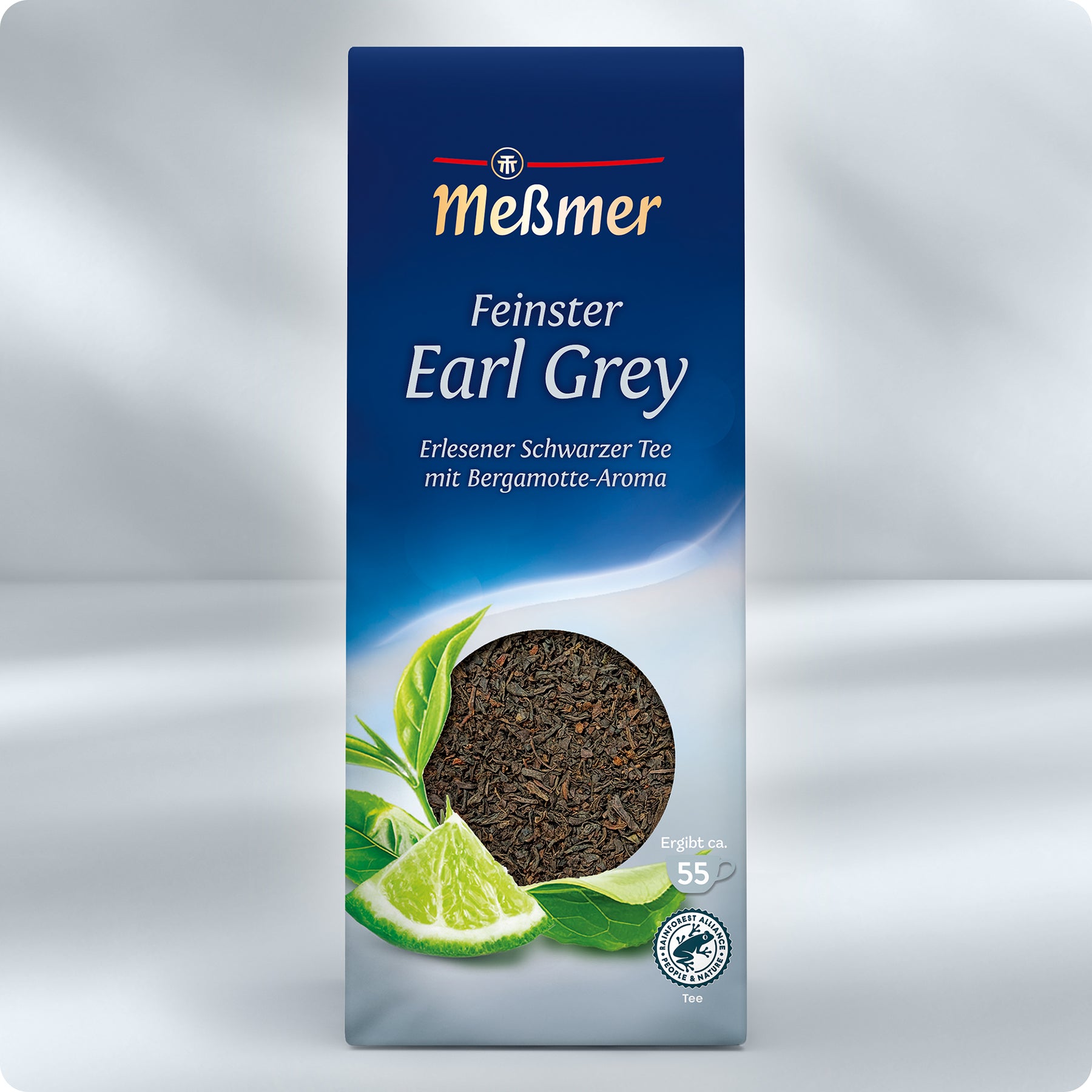 Eine Schachtel Meßmer DE Feinster Earl Grey Tee zeigt ein kreisrundes Fenster mit losem Schwarztee und Abbildungen einer Limettenscheibe, einer Bergamotte und grüner Blätter auf blauem und hellem Hintergrund.