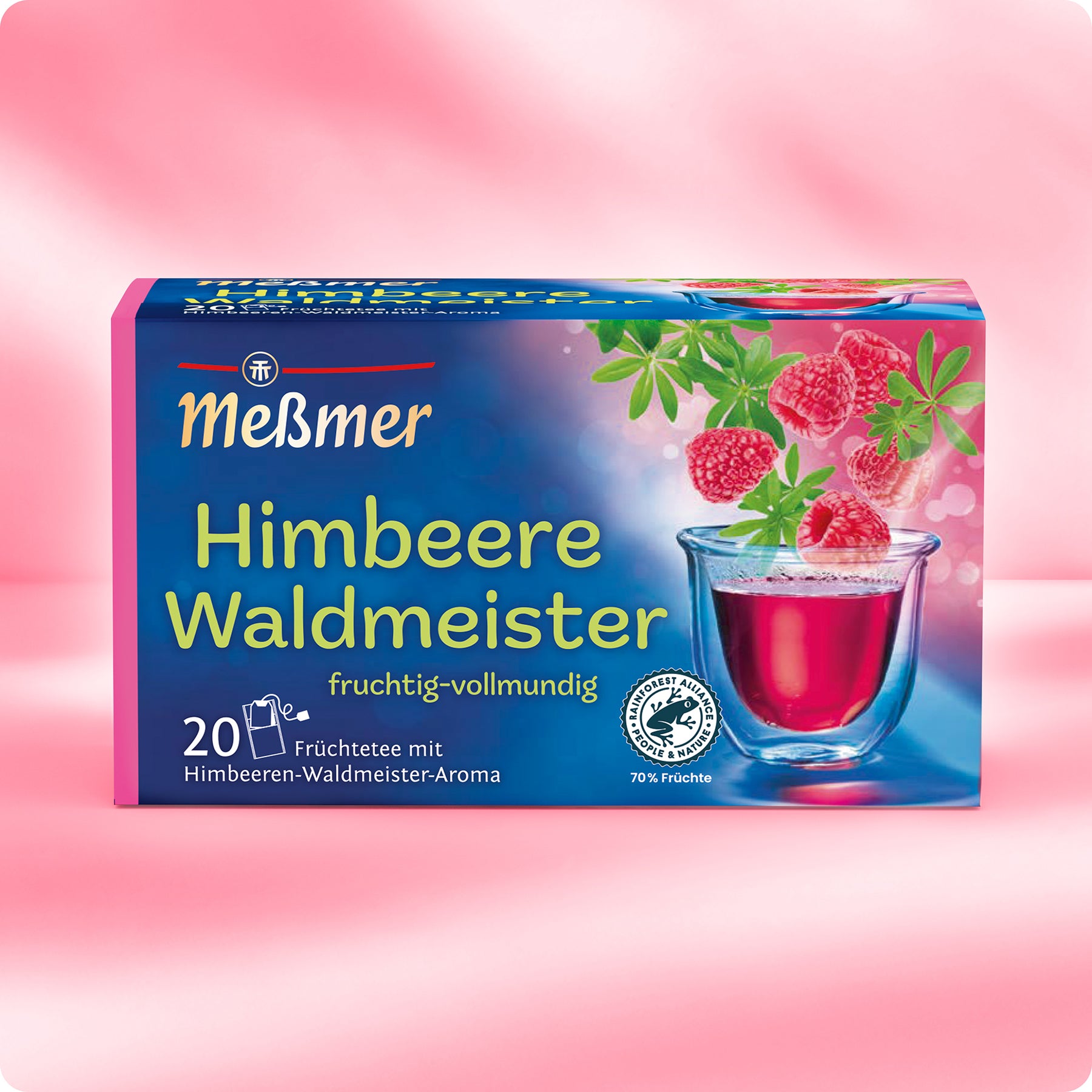 Eine Schachtel Meßmer DE Himbeere-Waldmeister-Tee mit Himbeeren, Waldmeisterblättern, rotem Tee und einem Verpackungstext mit 20 Teebeuteln, 70% Fruchtanteil und "Tee des Jahres" steht auf einem rosa Hintergrund.