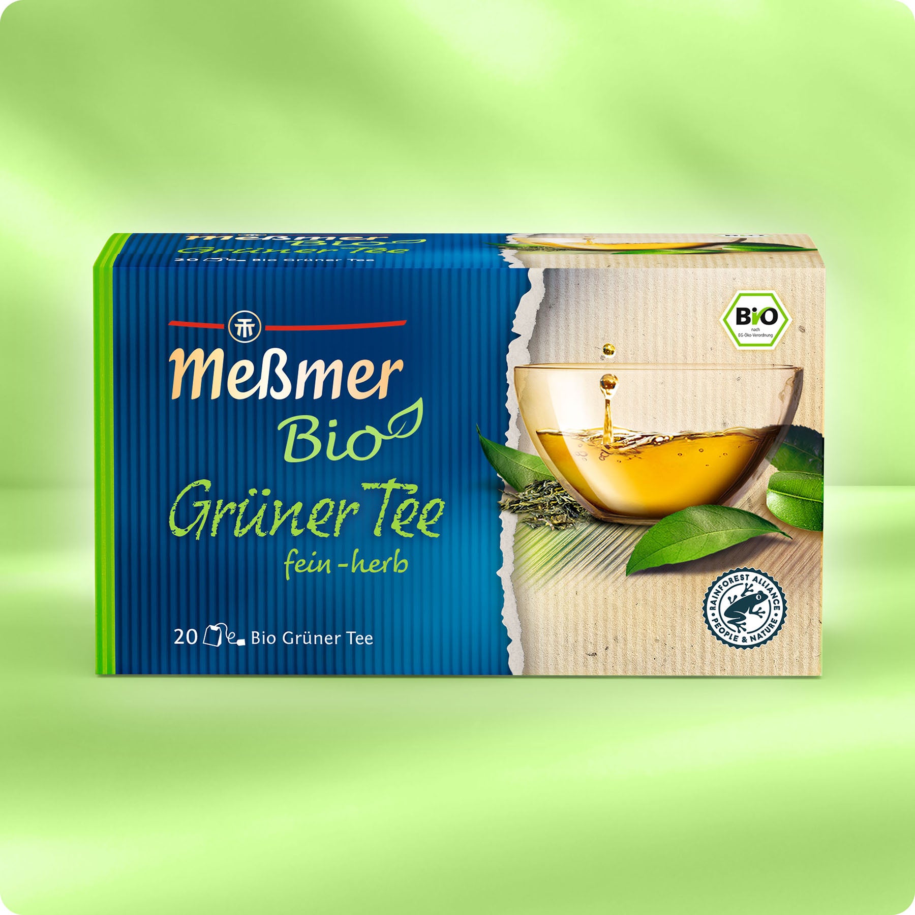 Neben frischen Teeblättern und einer Glastasse steht eine Schachtel Meßmer DE Bio Grüner Tee. Die grün-beigefarbene Verpackung auf hellgrünem Hintergrund ist ideal für alle Liebhaber von Bio-Grüntee, die den puren Geschmack von Meßmer suchen.