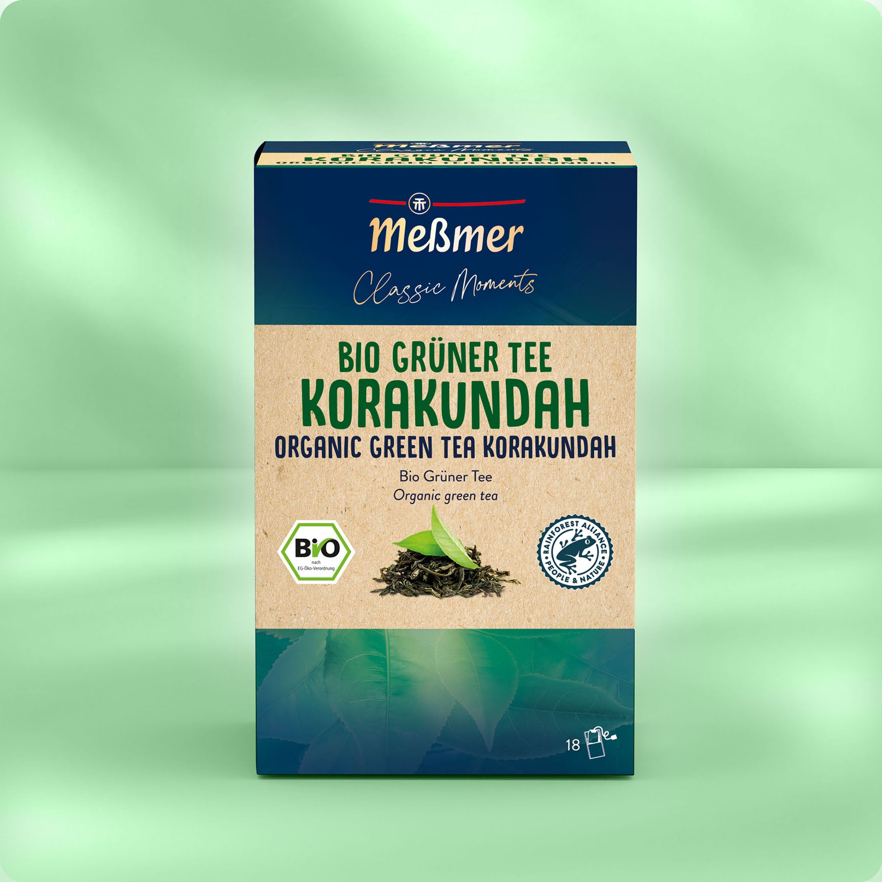 Eine Kiste Meßmer DE Bio Grüner Tee Korakundah steht aufrecht vor einem sanft verschwommenen grünen Hintergrund, mit deutschem und englischem Text sowie einem BIO-Zertifizierungsetikett auf der Verpackung.