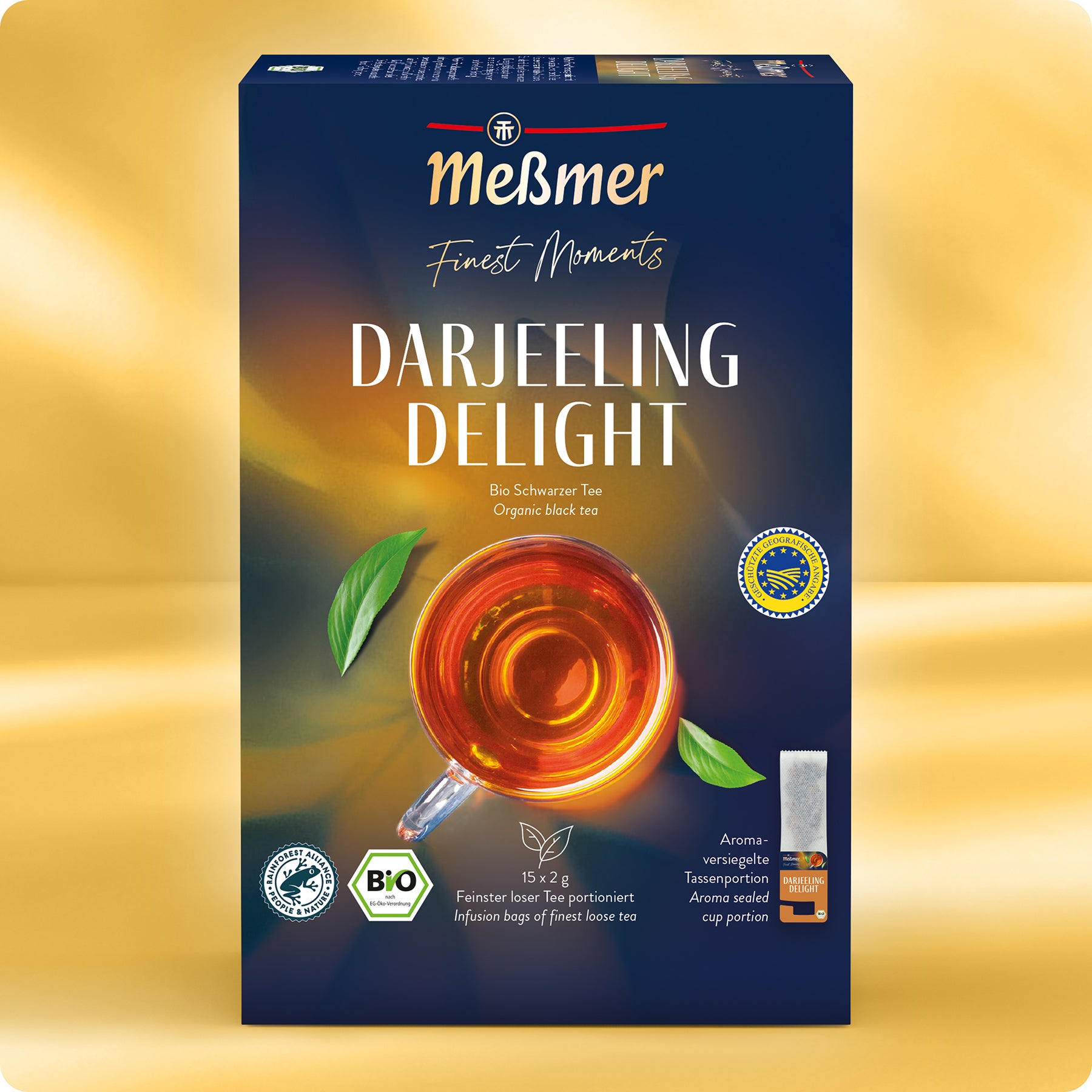 Schachtel Meßmer DE Bio Darjeeling Delight Bio-Schwarztee, abgebildet mit der Draufsicht einer bernsteinfarbenen Teetasse, Produktzertifizierungen, goldenen und blauen Akzenten sowie einem deutsch-englischen Text, der die Teetradition von Meßmer hervorhebt.