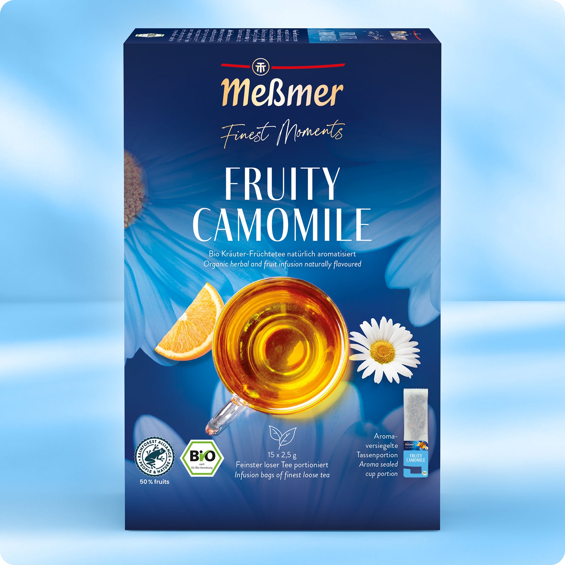 Eine Schachtel Meßmer DE Bio Fruchtiger Kamillentee, die eine durchsichtige Tasse mit goldenem Tee, Kamillenblüten, Orangenkeilen, blauer Blumengrafik und Bio-Tee-Logos zusammen mit den wichtigsten Produktangaben zeigt.