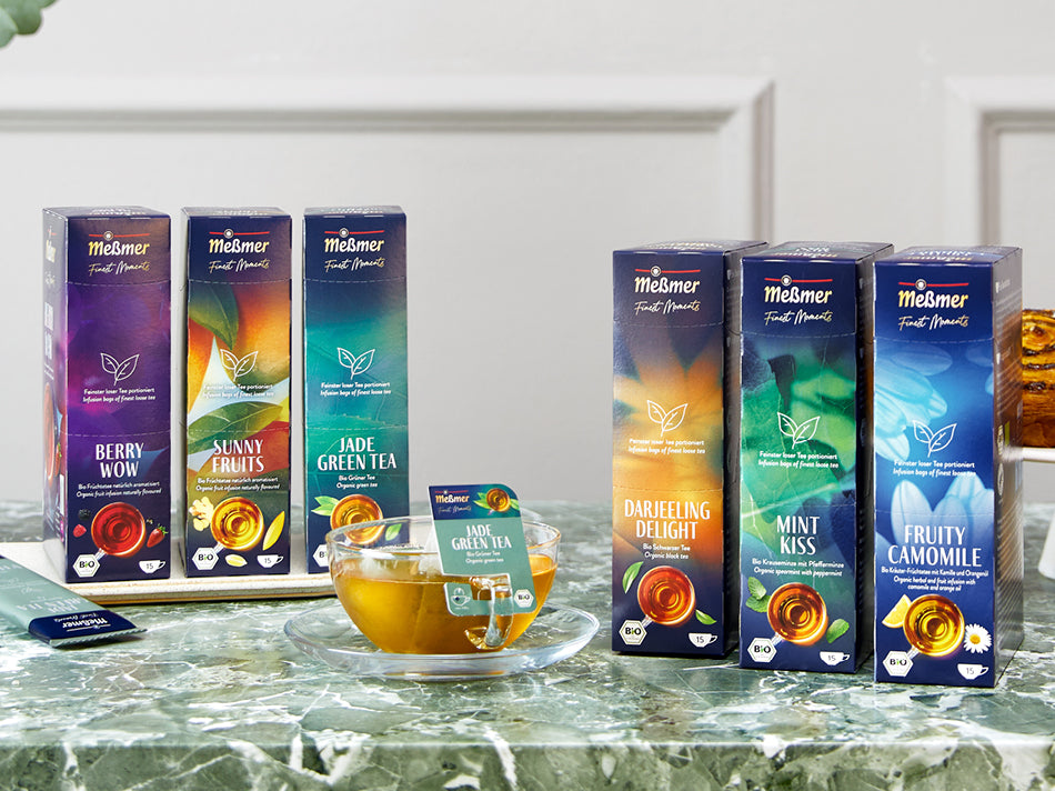Sechs Meßmer Teepackungen in verschiedenen Sorten wie Berry Wow, Sunny Fruits und Mint Kiss auf Marmoroberfläche, dazu Glas mit grünem Tee und Teebeutel darin.