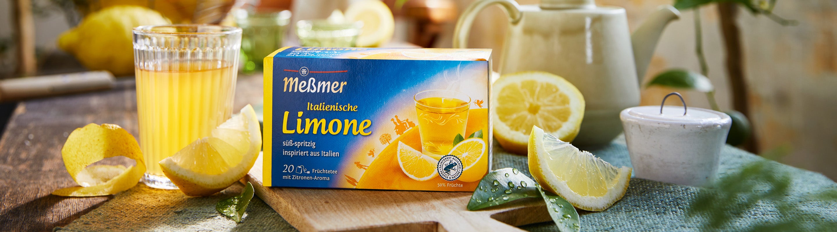 Eine Schachtel Meßmer Limone-Tee steht auf einem Holzbrett, umgeben von einem Glas Zitronentee, Zitronenscheiben, Zitronenschalen, einer Teekanne und einem kleinen Glas, alles auf einem sonnenbeschienenen Tisch.