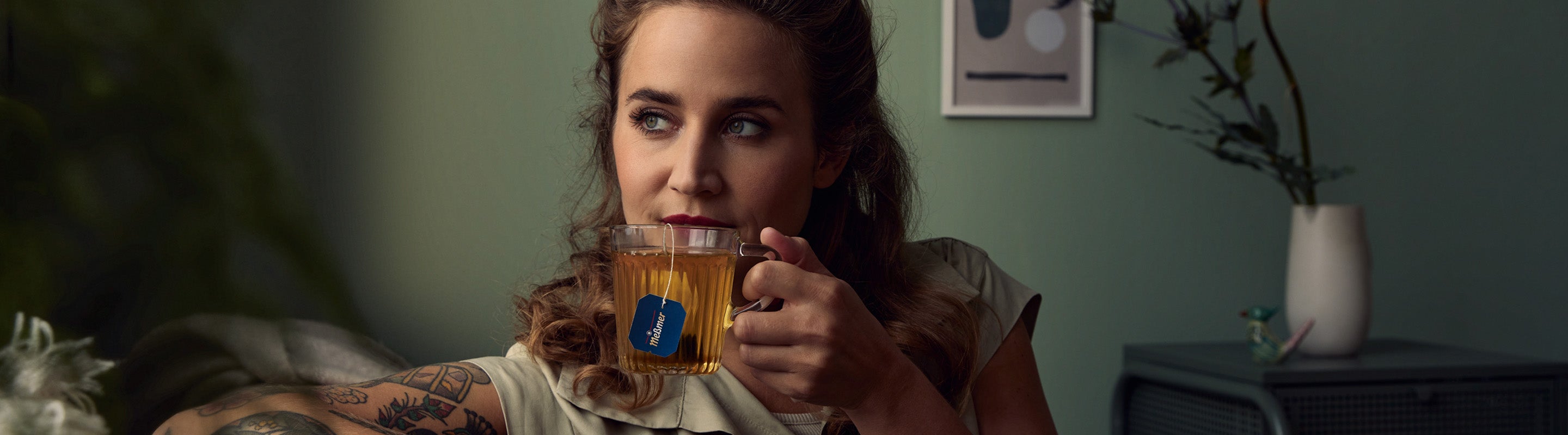 Eine Frau mit gewelltem Haar und Tattoos hält eine gläserne Teetasse und schaut nachdenklich zur Seite. Sie sitzt in einem sanft beleuchteten Raum mit grünen Wänden, einer Topfpflanze und gerahmter Kunst im Hintergrund.
