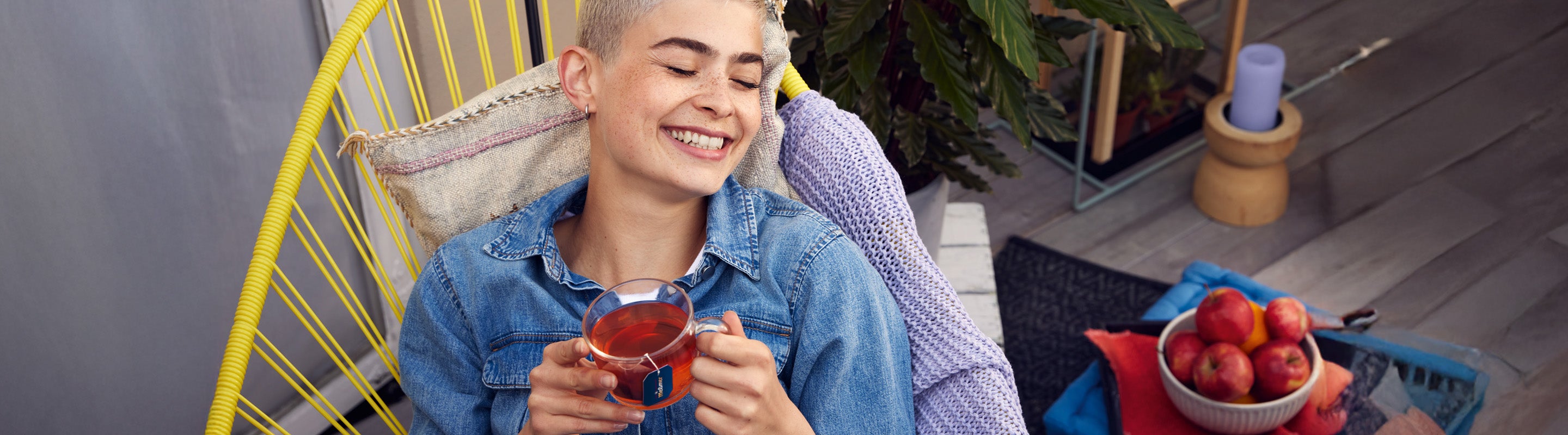 Eine Person mit kurzen Haaren lächelt mit geschlossenen Augen, hält eine Glastasse mit Tee in der Hand und entspannt sich in einem gelben Stuhl. Eine Schale mit Äpfeln und eine violette Kerze stehen auf einem Tisch in der Nähe.