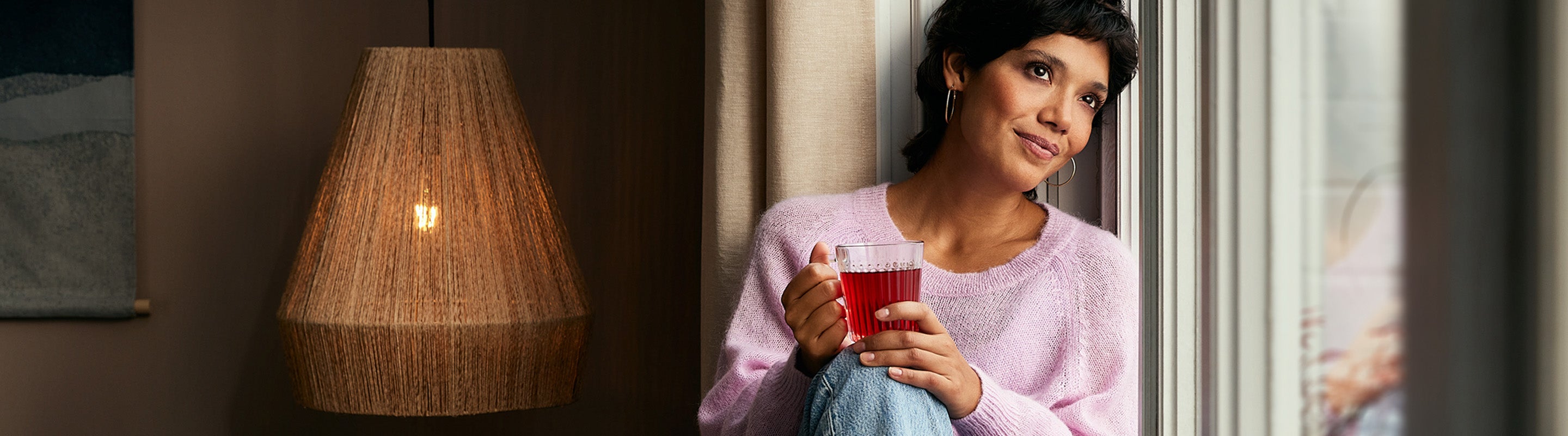 Eine Frau mit kurzen dunklen Haaren sitzt am Fenster, hält eine Glastasse mit rotem Tee in der Hand und trägt einen hellvioletten Pullover und Jeans. Sie blickt nachdenklich nach draußen. Neben ihr leuchtet eine warme, geflochtene Lampe.