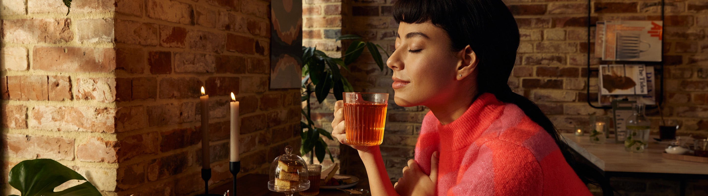 Eine Frau in einem rosafarbenen Pullover sitzt an einem gemütlichen Tisch mit Kerzen, Desserts und Pflanzen und hält eine Glastasse mit Tee an ihr Gesicht, während sie die Augen schließt und das Aroma genießt.